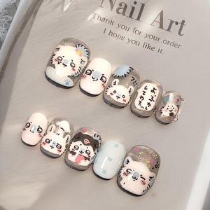 May include: A set of press-on nails with cartoon cat designs. The nails feature white, grey, and clear bases with hand-painted cat faces and Japanese text. The background includes the text "Nail Art" and a thank you message.