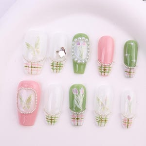 May include: A collection of ten artificial nails in various designs. The nails feature a mix of colors, including pink, green, and clear, with decorative elements such as butterflies, floral patterns, and pearl accents. Each nail has a plaid design at the base.