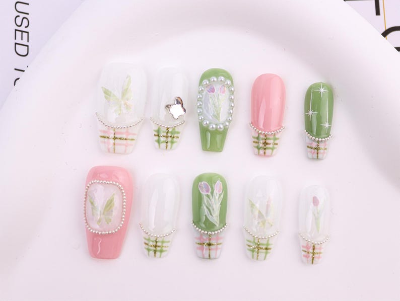 May include: A collection of ten artificial nails in various designs. The nails feature a mix of colors, including pink, green, and clear, with decorative elements such as butterflies, floral patterns, and pearl accents. Each nail has a plaid design at the base.