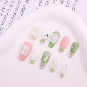 May include: A collection of artificial nails in various designs. Some nails are pink or green, adorned with floral patterns, butterflies, and rhinestones. Others feature a plaid design at the base. The nails are arranged on a white surface.