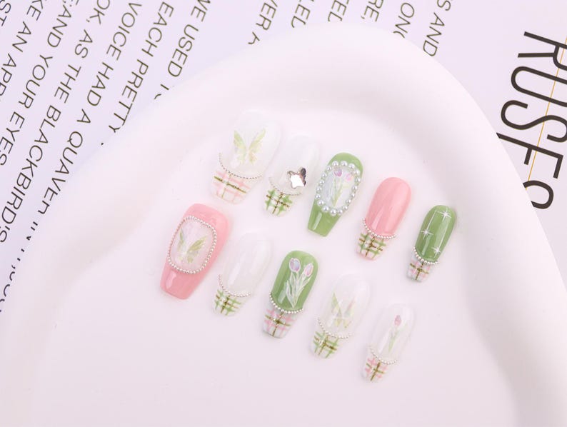 May include: A collection of artificial nails in various designs. Some nails are pink or green, adorned with floral patterns, butterflies, and rhinestones. Others feature a plaid design at the base. The nails are arranged on a white surface.