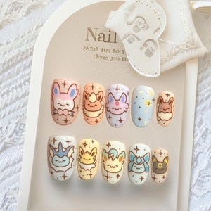 May include: A set of ten press-on nails featuring pastel-colored backgrounds and hand-painted illustrations of various cartoon creatures. Each nail is decorated with small star accents and distinct character faces, arranged on a display card.