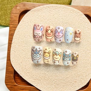 May include: A set of ten press-on nails featuring pastel-colored backgrounds and various cartoon character illustrations. Each nail is decorated with small star motifs and brown outlines. The designs include diverse creatures with pointed ears and expressive faces.