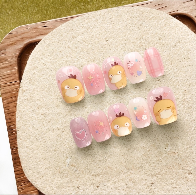 May include: A set of ten press-on nails featuring a light pink color palette with glossy finishes. The designs include illustrations of a yellow cartoon duck character, sparkling star accents, small white hearts, floral motifs, and a pink plaid pattern.