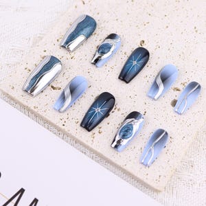 May include: Set of ten blue and silver press-on nails with various designs. The nails are arranged on a light beige surface.