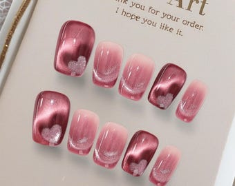 May include: A set of artificial nails in various shades of pink and burgundy, with a heart-shaped glitter design. The nails have a glossy finish and are arranged on a white card with the text "Nail Art".