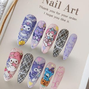 May include: A set of ten long, oval-shaped artificial nails with various cartoon character designs. The nails feature characters in purple, pink, and blue, with a plaid pattern and star designs. The text "Nail Art" and a thank you message are visible.