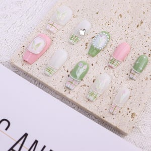 May include: A collection of artificial nails in various designs. The nails feature pastel pink, green, and white colors with floral and plaid patterns. Some nails are adorned with pearls and small embellishments.
