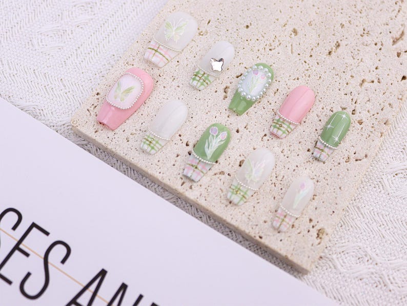 May include: A collection of artificial nails in various designs. The nails feature pastel pink, green, and white colors with floral and plaid patterns. Some nails are adorned with pearls and small embellishments.