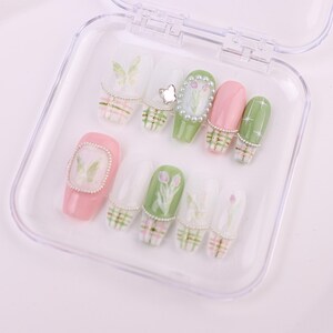 May include: A set of artificial nails in a clear plastic case. The nails feature various designs, including butterflies, flowers, and plaid patterns. Colors include pink, green, and white, with pearl accents. The nails are designed for a manicure.