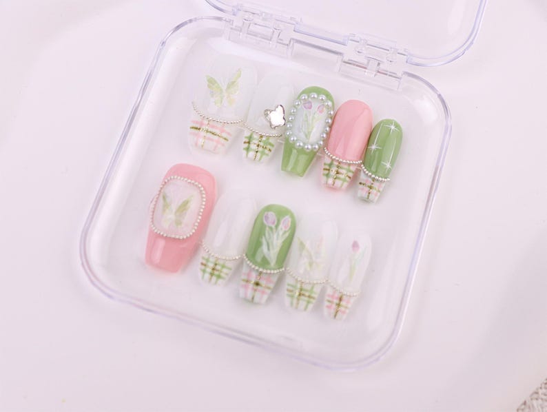 May include: A set of artificial nails in a clear plastic case. The nails feature various designs, including butterflies, flowers, and plaid patterns. Colors include pink, green, and white, with pearl accents. The nails are designed for a manicure.
