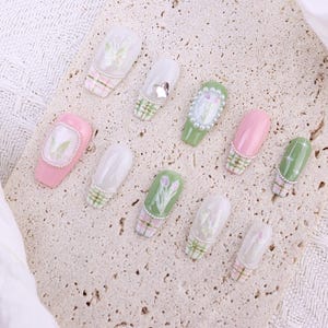May include: A collection of artificial nails with various designs. Some nails are pink or green, others are clear with floral patterns. Several nails feature a plaid design at the base, and some are adorned with pearls or small embellishments. The nails are arranged on a textured surface.
