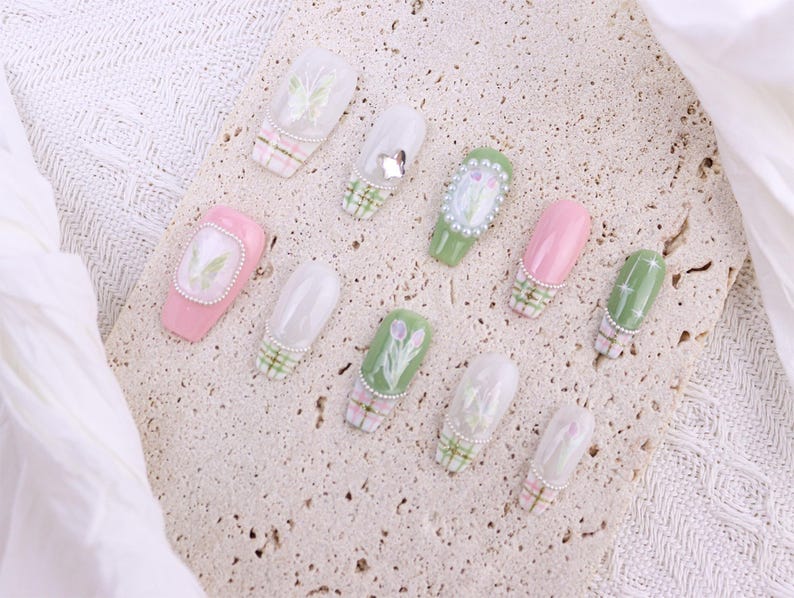 May include: A collection of artificial nails with various designs. Some nails are pink or green, others are clear with floral patterns. Several nails feature a plaid design at the base, and some are adorned with pearls or small embellishments. The nails are arranged on a textured surface.