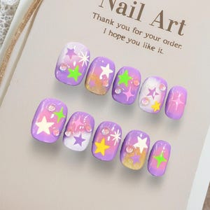 May include: Set of ten artificial nails with a purple gradient design, decorated with stars in white, yellow, and green, and small rhinestones. The nails are arranged on a neutral-colored card with the text "Nail Art".