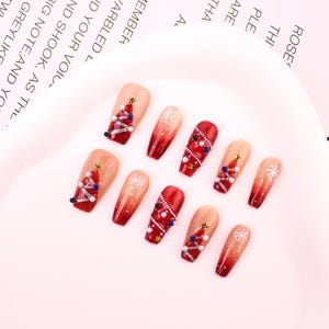Puede incluir: Un conjunto de diez uñas postizas con temática navideña. Las uñas presentan un diseño degradado de nude a rojo, con decoraciones de árboles de Navidad, copos de nieve y detalles de perlas y gemas.