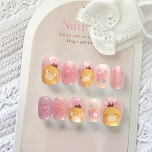 May include: A set of ten press-on nails arranged on a display card with a white bow. The nails feature a light pink base with various designs including a yellow cartoon duck character, glittery stars, small hearts, and a plaid pattern on one nail.