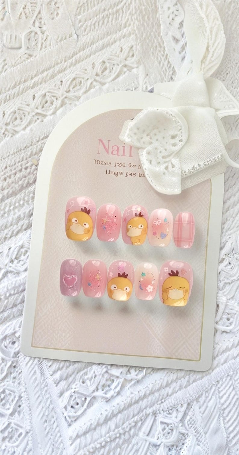 May include: A set of ten press-on nails arranged on a display card with a white bow. The nails feature a light pink base with various designs including a yellow cartoon duck character, glittery stars, small hearts, and a plaid pattern on one nail.