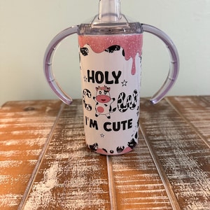 May include: A pink and white stainless steel tumbler with two handles and a clear lid. The tumbler has a cow graphic and the text "HOLY COW I'M CUTE".