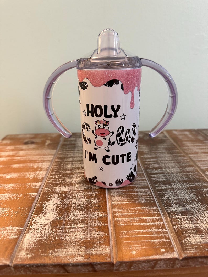 May include: A pink and white stainless steel tumbler with two handles and a clear lid. The tumbler has a cow graphic and the text "HOLY COW I'M CUTE".
