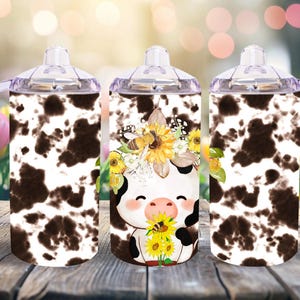 May include: Three stainless steel tumblers with a cow print design and a cute cartoon cow with a sunflower crown. The tumblers have clear lids and handles.