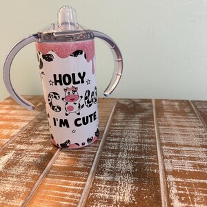 May include: A pink and white stainless steel tumbler with a cow design and the text "HOLY COW I'M CUTE". The tumbler has two handles and a clear lid with a straw.