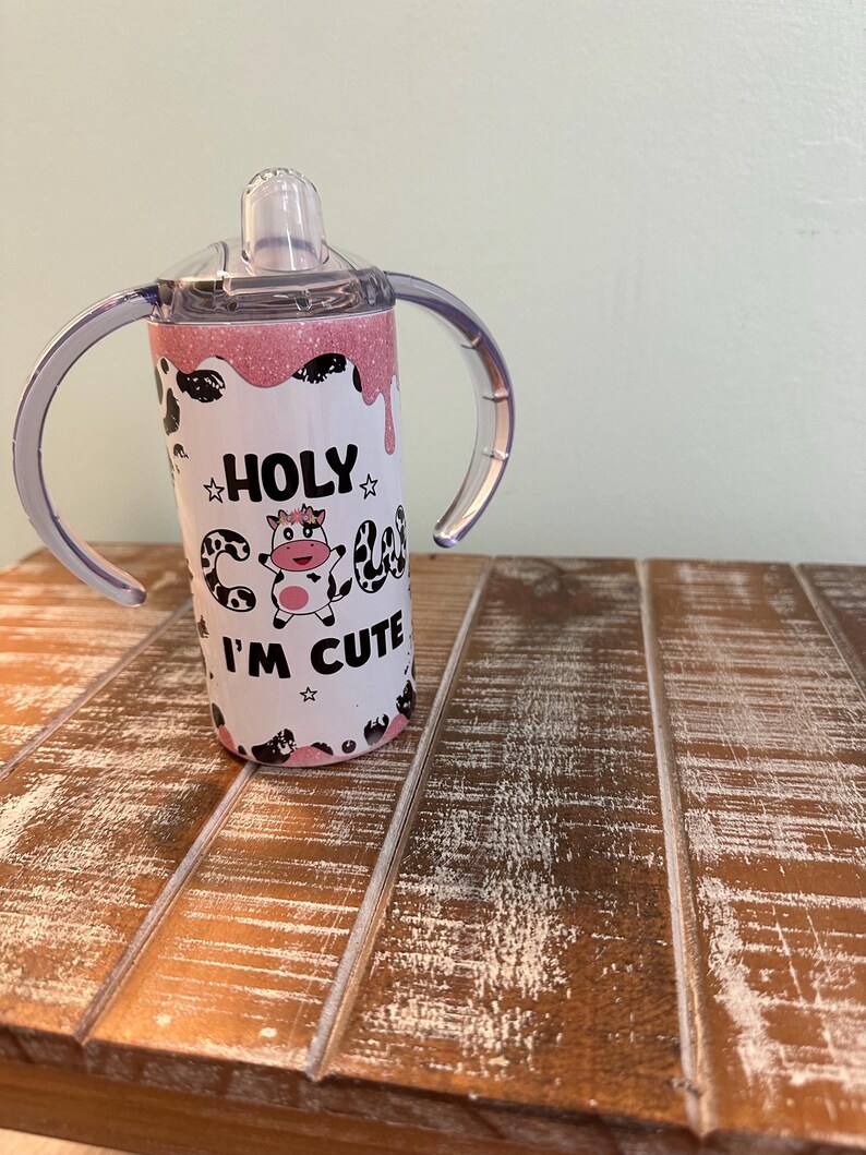 May include: A pink and white stainless steel tumbler with a cow design and the text "HOLY COW I'M CUTE". The tumbler has two handles and a clear lid with a straw.