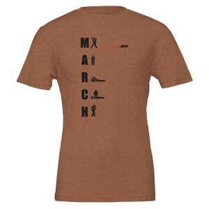Op de afbeelding: Een bruin t-shirt met het woord "MARCH" verticaal gedrukt in zwarte blokletters. Elke letter wordt vergezeld door een kleine, zwarte illustratie van een stokfiguur die verschillende activiteiten uitbeeldt, zoals het dragen van een tas of zitten.
