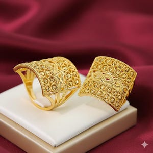 May include: Two gold-colored rings with intricate, textured designs. The rings have a wide, rectangular face with a detailed pattern of small beads and interwoven lines. The rings are displayed on a white surface against a burgundy background.