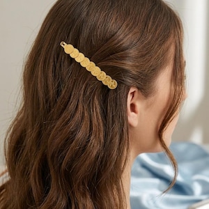 May include: A gold-toned hair clip featuring a linear arrangement of nine small, circular coin-like charms. Each charm displays a detailed embossed profile design. The hair accessory is secured horizontally, holding back a section of wavy brown hair.
