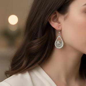 May include: Silver teardrop earrings with a detailed circular design in the center, surrounded by small, sparkling stones. The earrings hang from a simple hook, showcasing a classic and elegant jewelry style.