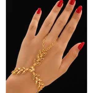 May include: A gold-colored hand chain bracelet featuring a floral and leaf design. The bracelet connects to a ring with a delicate chain. The jewelry is worn on a hand with red nail polish against a black background.