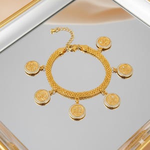 May include: Gold-coloured bracelet with a chain-link design and coin charms. Each charm has a detailed design and is accented with small, clear crystals. The bracelet is displayed on a reflective surface.