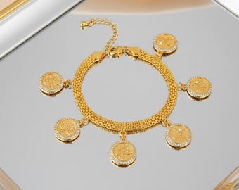 May include: Gold-toned bracelet with a chain-link design and dangling coin charms. Each charm features a detailed design and is accented with small, clear crystals. The bracelet is displayed on a reflective surface.