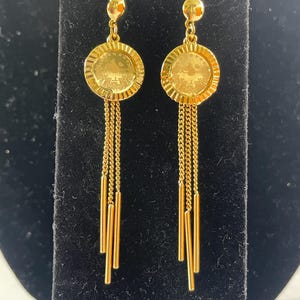 Op de afbeelding: Een paar goudkleurige oorhangers. Elke oorbel heeft een cirkelvormige hanger met een gedetailleerd ontwerp, opgehangen aan een haak. Onder de hanger vallen fijne kettingen en gouden staven in een waterval naar beneden, wat een vloeiend effect creëert. De oorbellen worden tegen een zwarte achtergrond gepresenteerd.