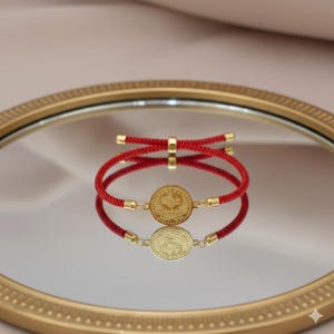 May include: Red cord bracelet with a gold-colored coin pendant. The coin features a detailed design and is connected to the cord with gold-colored accents. The bracelet is displayed on a reflective surface.