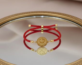 May include: Red cord bracelet with a gold-colored coin pendant. The coin features a detailed design and is connected to the cord with gold-colored accents. The bracelet is displayed on a reflective surface.