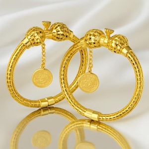 May include: Two gold-colored bangles with a textured design. Each bangle features a decorative sphere and a small gold coin pendant hanging from a chain. The bangles are displayed on a reflective surface.