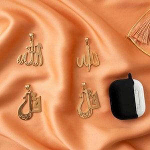 May include: Four gold-coloured metal pendants featuring intricate Arabic calligraphy designs, each with a small top loop for attachment. Next to the pendants is a black and white wireless earbud charging case with a smooth, rounded rectangular shape.