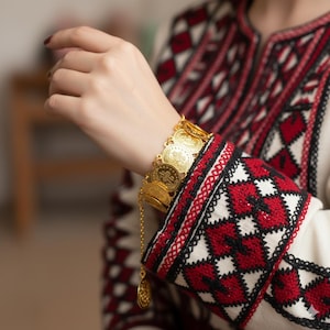 Pode incluir: Uma pulseira dourada com moedas e um pingente pendurado é usada no pulso. A pulseira é feita de várias moedas circulares. A manga de uma peça de roupa branca com bordados vermelhos e pretos é visível.