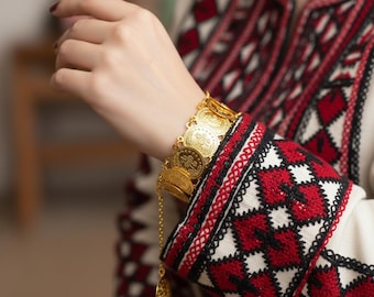 May include: A gold-colored coin bracelet with a dangling charm is worn on a wrist. The bracelet is made of multiple circular coins. The sleeve of a white garment with red and black embroidery is visible.