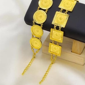 May include: Two gold-coloured bracelets. One features round coin-like charms, the other square charms. Both have intricate designs and chain extensions. The bracelets are displayed on a black surface against a white background.