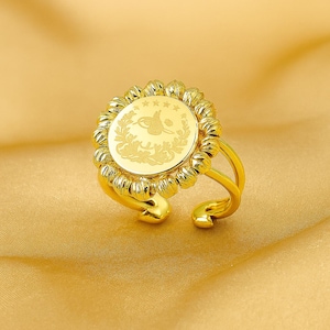 May include: A gold-colored ring with a circular centerpiece featuring an engraved design. The ring has a floral-like border around the center and a split band. The ring is displayed on a gold-colored fabric.