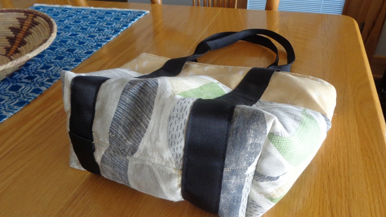 May include: A beige and gray tote bag with black straps. The bag has a patchwork design with different patterns and colors. The bag is made of a soft, fabric material.