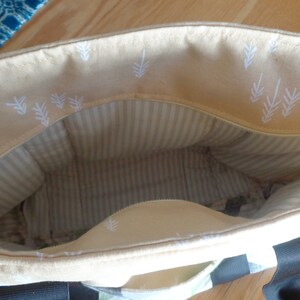May include: A beige tote bag with a zipper closure and white leaf pattern on the inside. The bag has a striped lining and black handles.