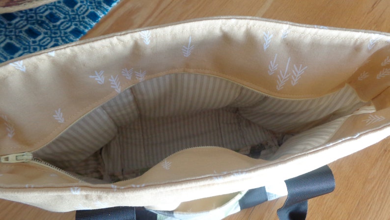 May include: A beige tote bag with a zipper closure and white leaf pattern on the inside. The bag has a striped lining and black handles.
