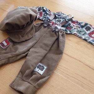 May include: A brown corduroy outfit for a baby or toddler, featuring a cap, pants, and a shirt with a patchwork design of Christmas scenes. The pants have a pair of suspenders and decorative patches.