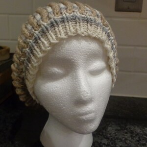 May include: A knitted hat with a beige, gray, and white color pattern. The hat is made of soft yarn and has a ribbed texture.