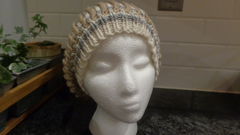 May include: A knitted hat with a beige, gray, and white color pattern. The hat is made of soft yarn and has a ribbed texture.