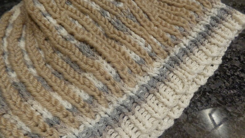 May include: A close-up of a knitted hat with a beige, white, and gray striped pattern.