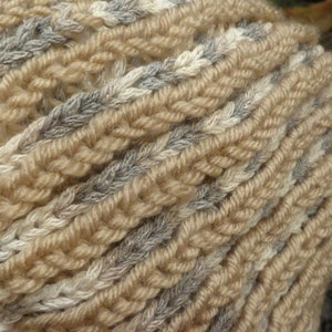 May include: A close-up of a knitted scarf in beige, white, and gray. The scarf is made of a soft, textured yarn and has a braided pattern.